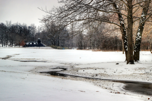 A snowy landscape with a winding stream, bare trees, and a distant building in a serene winter setting.