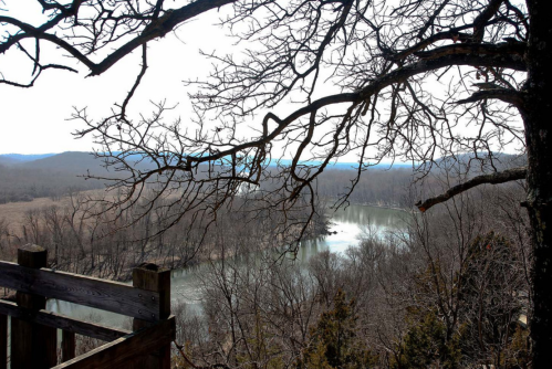 A scenic view of a river surrounded by bare trees and rolling hills under a cloudy sky.