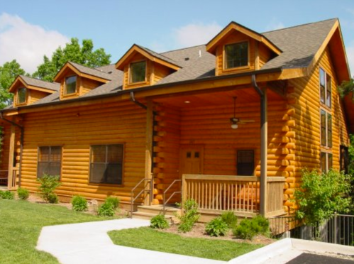 A large log cabin with a wooden porch, multiple windows, and a green lawn under a clear blue sky.