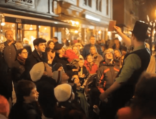A street performer entertains a captivated crowd at night, with people of all ages watching intently.