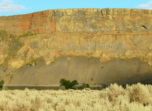 A rugged cliff with colorful layers rises above a flat, dry landscape dotted with sparse vegetation.