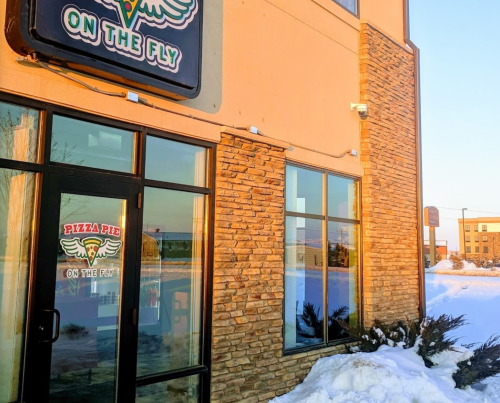 Exterior of "Pizza Pie on the Fly" restaurant, featuring a stone facade and snow-covered ground.