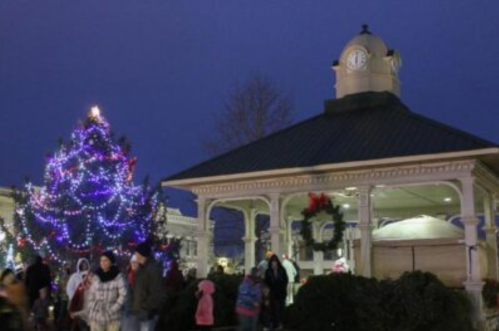 A festive scene with a decorated Christmas tree and a gazebo, surrounded by people in winter clothing at dusk.