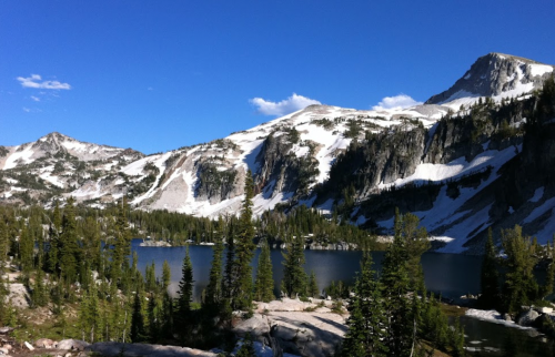A serene mountain landscape featuring a lake surrounded by snow-capped peaks and lush green trees under a clear blue sky.