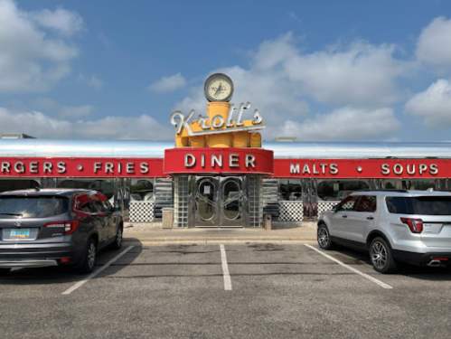 A retro diner with a red and silver exterior, featuring a sign that reads "Kroll's Diner" and parked cars in front.