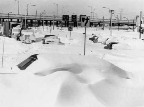 A snowy scene with buried cars and a highway sign, depicting a heavy snowstorm in a city.