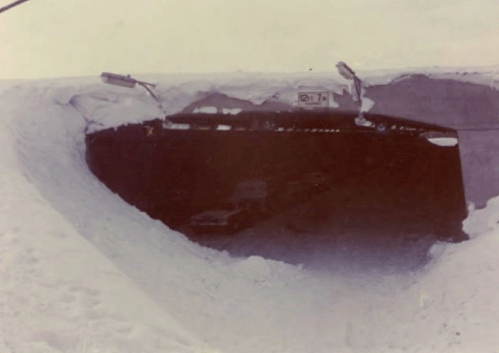 A large snowbank surrounds an opening, revealing a dark space with vehicles inside, suggesting a snow-covered area.
