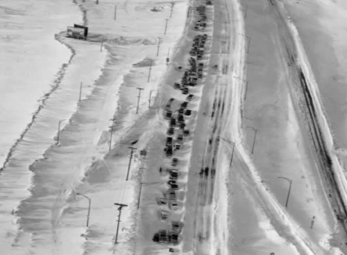 Aerial view of a long line of cars stuck in snow on a highway, surrounded by snow-covered fields.