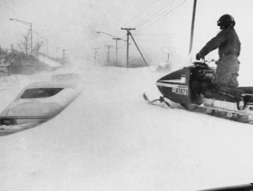 A snowmobiler navigates through deep snow, with a buried car visible in the background during a blizzard.