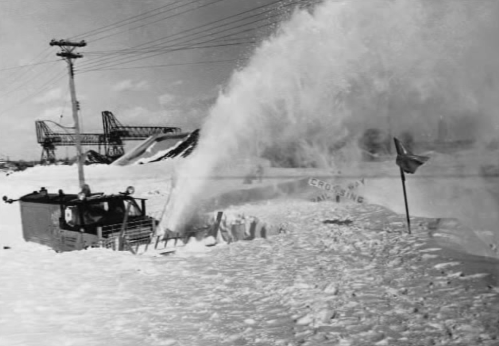 A snowplow clears a snowy landscape, with snow blowing and power lines in the background. Black and white image.