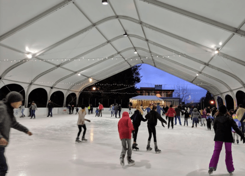 A lively ice skating rink under a large tent, with people skating and festive lights in the evening sky.