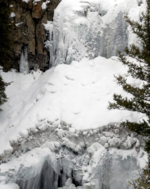 A frozen waterfall surrounded by snow-covered rocks and evergreen trees.