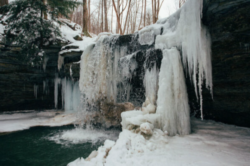A frozen waterfall surrounded by ice and snow, with a small pool of water at the base, set in a winter forest.