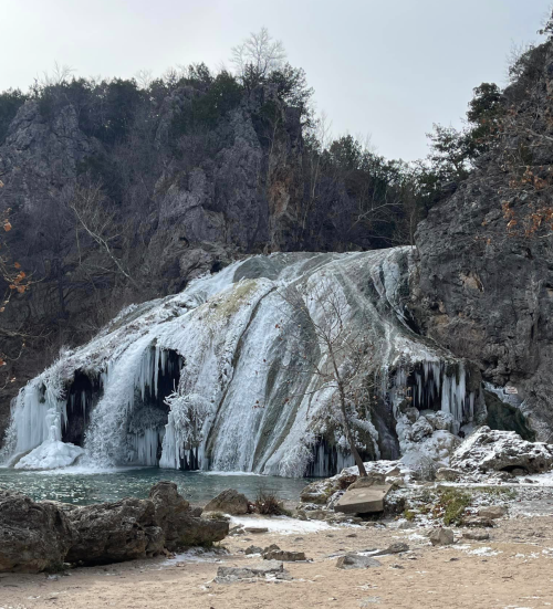 A frozen waterfall cascading down rocky cliffs, surrounded by snow and bare trees in a winter landscape.