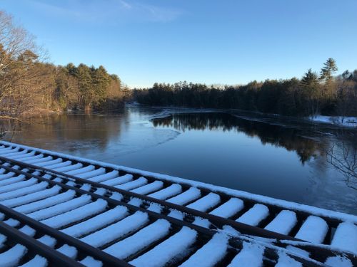 A serene winter scene featuring a snow-covered railway track beside a calm, icy river surrounded by trees.
