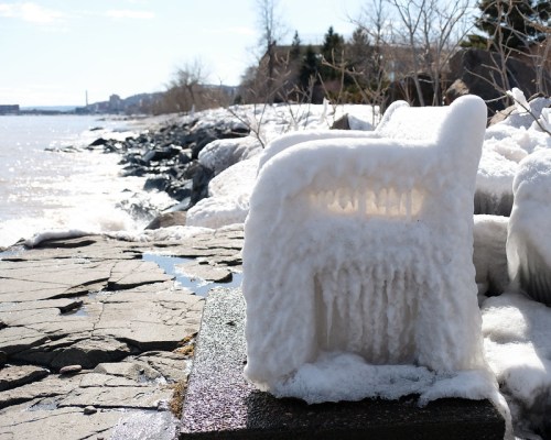 A frozen sculpture resembling a chair, covered in ice, near a rocky shoreline with water in the background.