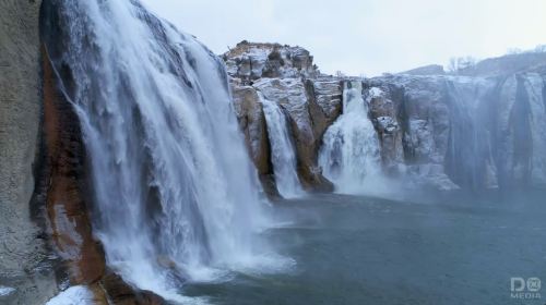 A scenic view of cascading waterfalls surrounded by snow-covered rocks and a misty river below.