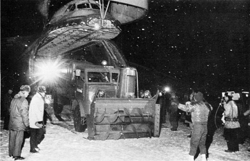 A snowplow truck is being unloaded from a large aircraft in a snowy environment, with people observing the operation.