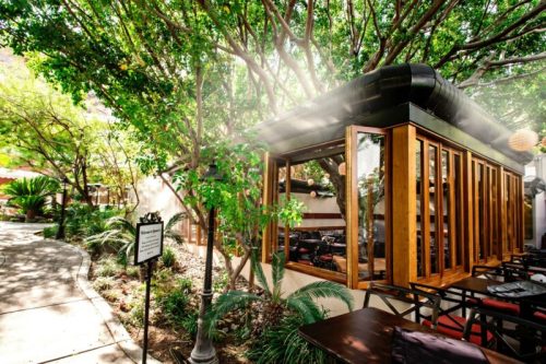 A cozy outdoor dining area surrounded by lush greenery and trees, featuring wooden structures and misting for comfort.
