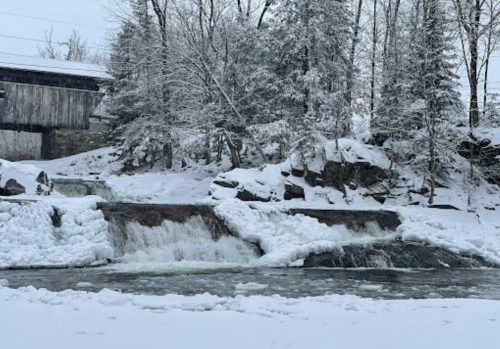 A snowy landscape featuring a frozen waterfall and a covered bridge in the background, surrounded by trees.