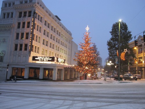 A snowy street scene featuring a brightly lit Christmas tree and a historic building with festive decorations.