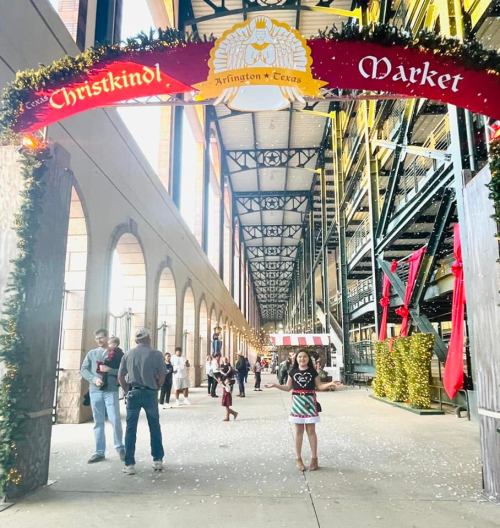 A festive market entrance with decorations, people walking, and a girl in a colorful outfit standing in the foreground.