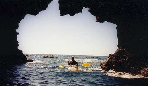 A kayaker paddles through a rocky archway, with the ocean and boats visible in the background under a clear sky.