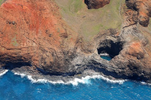 Aerial view of rugged coastline with red cliffs, green patches, and waves crashing against rocky formations.