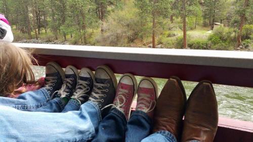 A row of feet in various shoes resting on a boat railing, with a scenic river and trees in the background.
