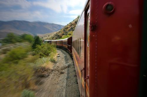 A vintage train curves along a scenic mountain track, surrounded by greenery and hills under a blue sky.
