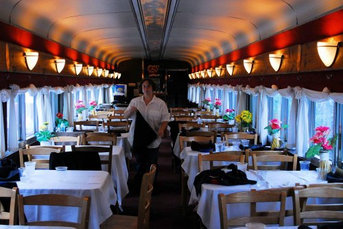 A dining car interior with tables set for meals, flowers on tables, and a server preparing for guests.