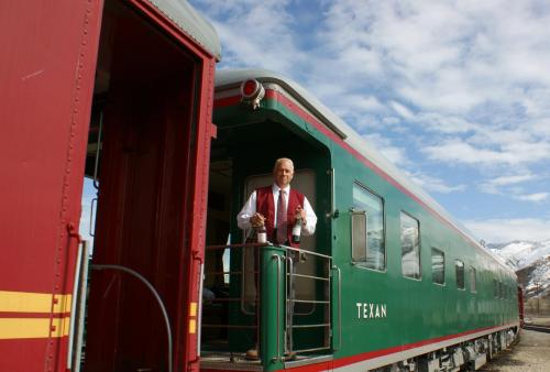 A man in a vest stands on the platform of a green train car against a backdrop of mountains and blue sky.
