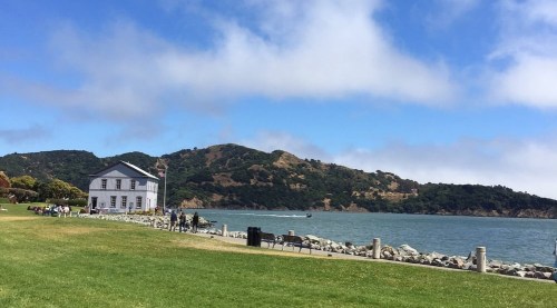 A scenic view of a waterfront with a white building, grassy area, and hills in the background under a blue sky.