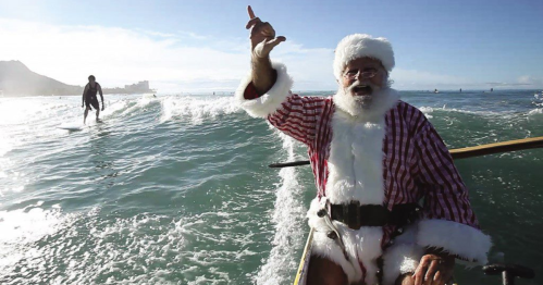 Santa Claus in a red and white outfit waves from a boat, with surfers and ocean waves in the background.