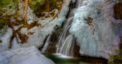 A serene waterfall cascading over icy rocks, surrounded by snow and lush green trees in a tranquil forest setting.