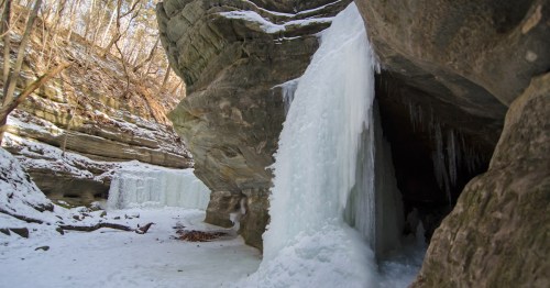 A frozen waterfall cascading over rocky cliffs, surrounded by snow-covered trees and a serene winter landscape.