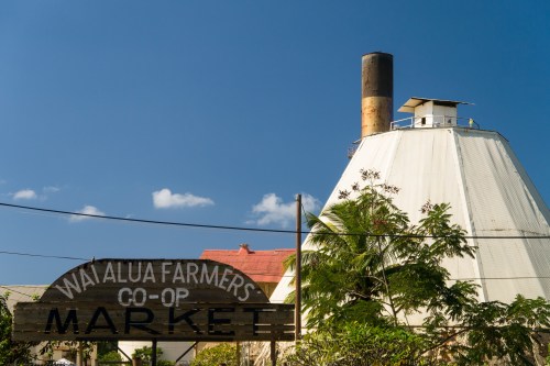Sign for Wai Alua Farmers Co-Op Market in front of a large, white, conical building against a blue sky.