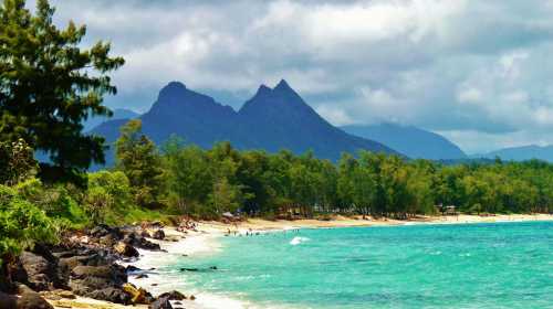 A serene beach with turquoise water, rocky shore, and lush green trees, set against a backdrop of mountains under cloudy skies.