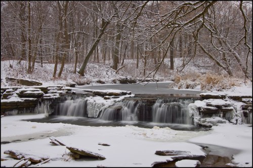 A serene winter scene featuring a frozen waterfall surrounded by snow-covered trees and a still, icy river.