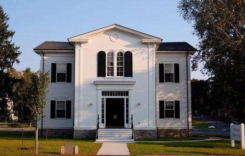 A large white two-story house with black shutters and a central entrance, surrounded by green grass and trees.
