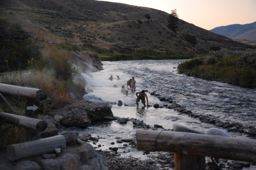 People enjoying a natural hot spring by a river, surrounded by hills and steam rising in the evening light.