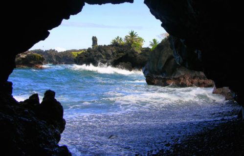 View of a rocky coastline through a cave opening, with waves crashing on the shore and greenery in the background.