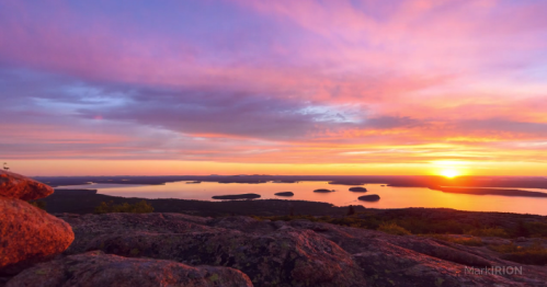 A vibrant sunset over a calm lake, with rocky foreground and scattered islands in the distance.