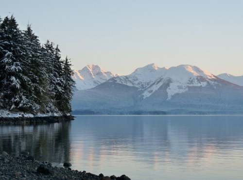 Serene landscape of snow-capped mountains reflecting in calm water, framed by evergreen trees along the shore.