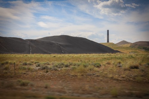 A landscape featuring rolling hills, sparse vegetation, and a tall industrial smokestack against a blue sky.