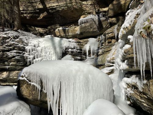A frozen waterfall with large icicles hanging from rocky cliffs, surrounded by snow-covered boulders and trees.