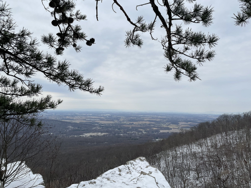 A winter landscape view from a rocky overlook, framed by pine branches, with a cloudy sky and distant hills.