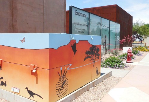 Colorful mural on a utility box with desert scenes, near an art gallery entrance surrounded by flowers and plants.