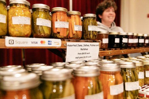 A display of jars filled with assorted jams, jellies, pickles, and relishes, with a smiling vendor in the background.