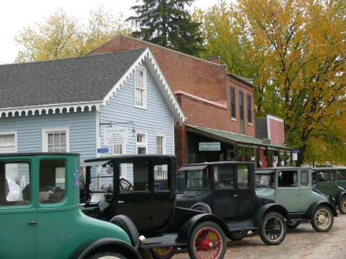A row of vintage cars parked in front of historic buildings with autumn trees in the background.